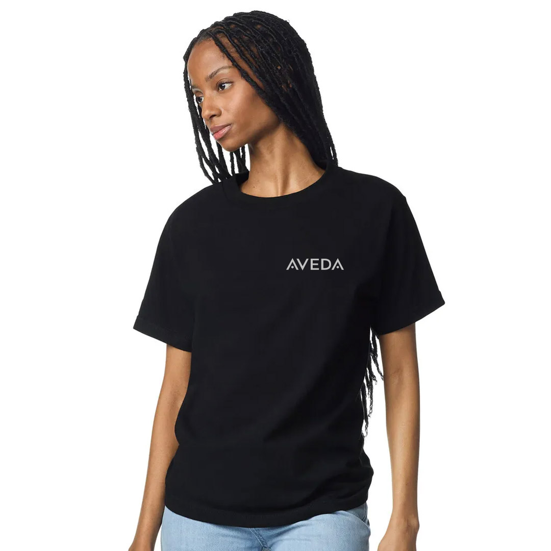 Vrou met 'n swart T-hemp met "AVEDA" op die linkerbors gedruk, staan en kyk na die kant met haar hande by haar heupe. | Imaginal Marketing Store - An Aveda Partner