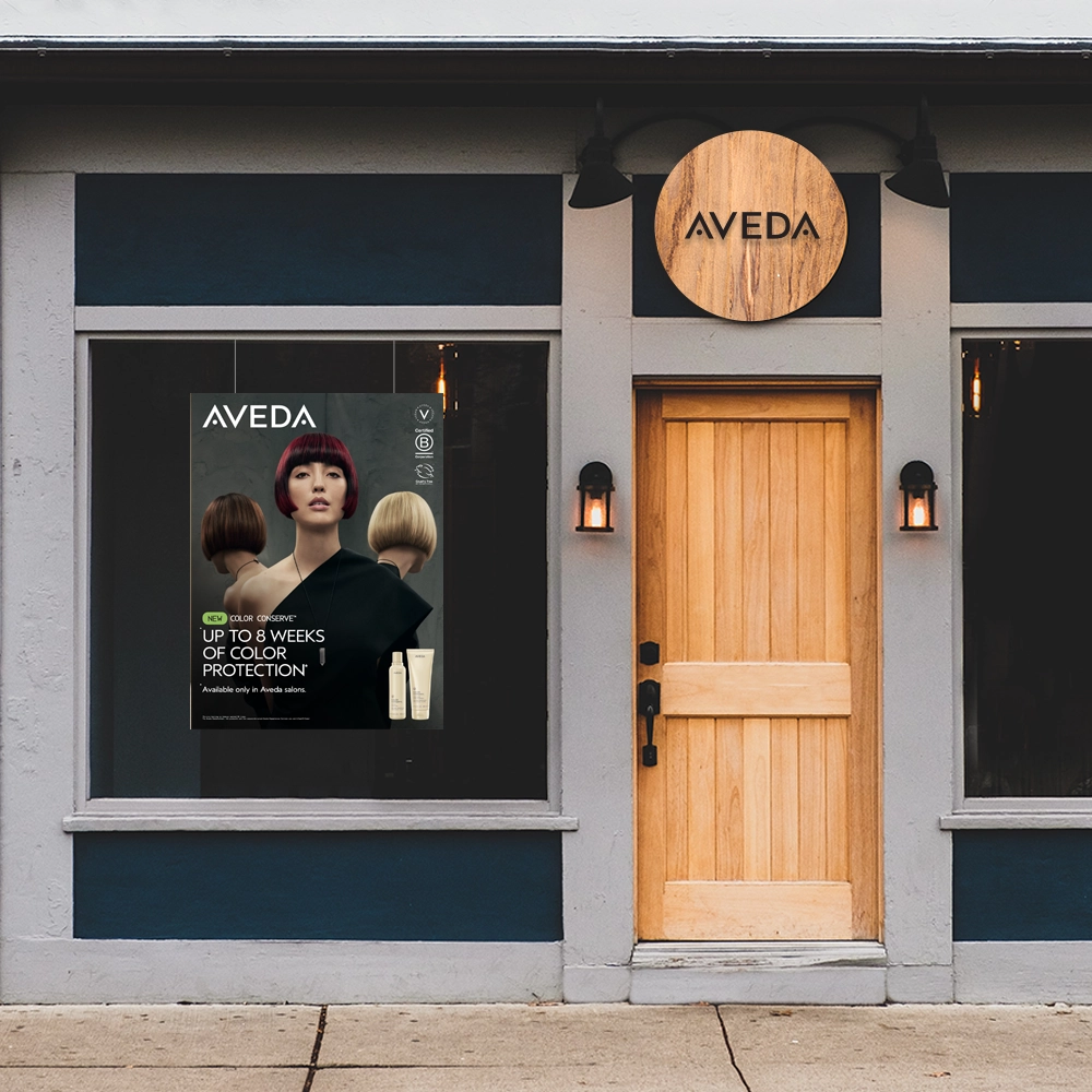 'n Winkelvenster met 'n houtdeur en twee vensters, een met 'n Aveda-plakkaat met haarmodelle en 'n produkadvertensie. | Imaginal Marketing Store - An Aveda Partner