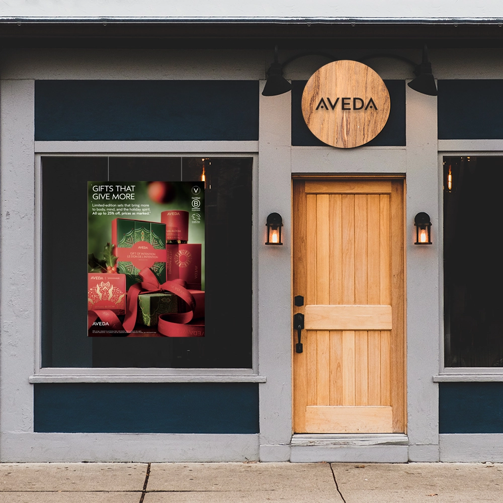 Winkelvenster met 'n houtdeur, twee muurligte en 'n AVEDA-teken bo. 'n Plakkaat met rooi geskenkbokse en die teks “GESKENKE WAT MEER GEE” word in die venster vertoon. | Imaginal Marketing Store - An Aveda Partner