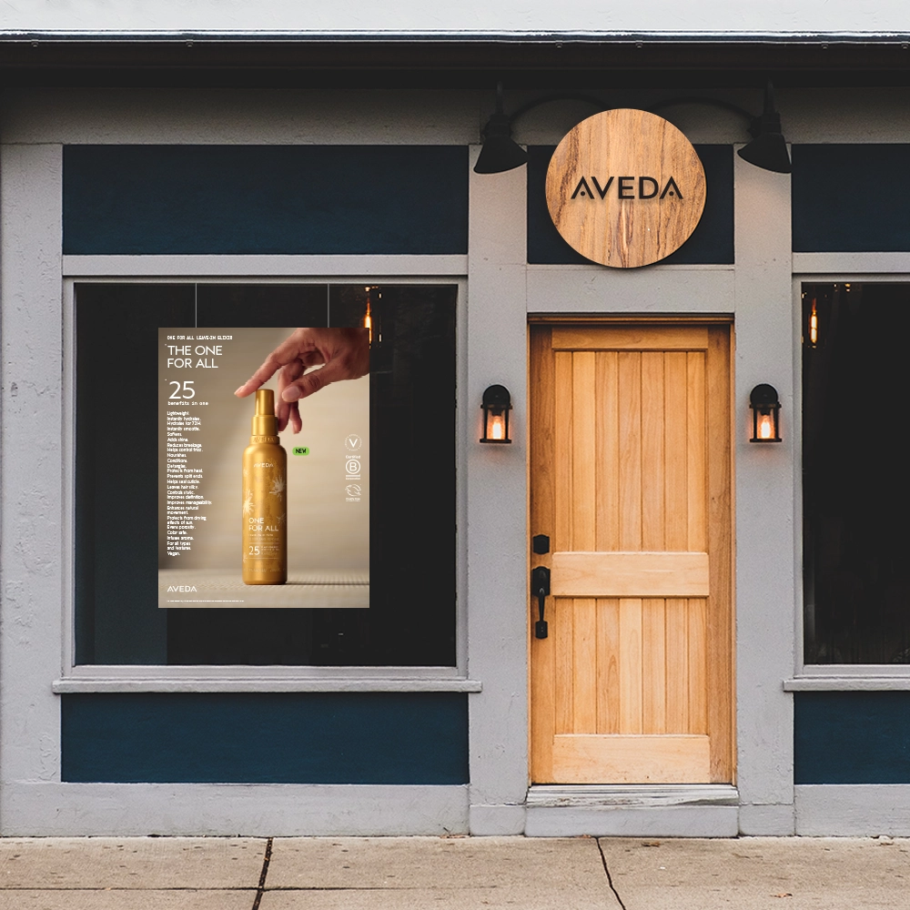 'n Winkelvenster met 'n houtdeur en 'n “Aveda”-teken, met 'n vensterplakkaat wat 'n goue bottel haarproduk adverteer en 'n hand wat die spuitkop druk. | Imaginal Marketing Store - An Aveda Partner